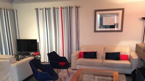 Un salon avec un canapé beige, deux fauteuils, une table basse, un téléviseur et des rideaux à rayures.