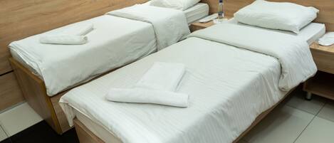 A hotel room with two single beds, a wooden headboard, a bedside table with a phone and a bottle, and a tiled floor.