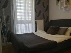 A bedroom with a bed, a radiator, sheer curtains, and wall-mounted shelves with potted plants.