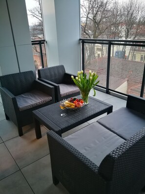 A balcony with wicker furniture, a table with fruit and a glass of wine, and a view of buildings and trees.