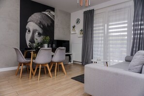 A modern living room with a grey sofa, a dining table with chairs, a large abstract painting, and a window with curtains. - Nova Apartments Rakowicka (Kraków)