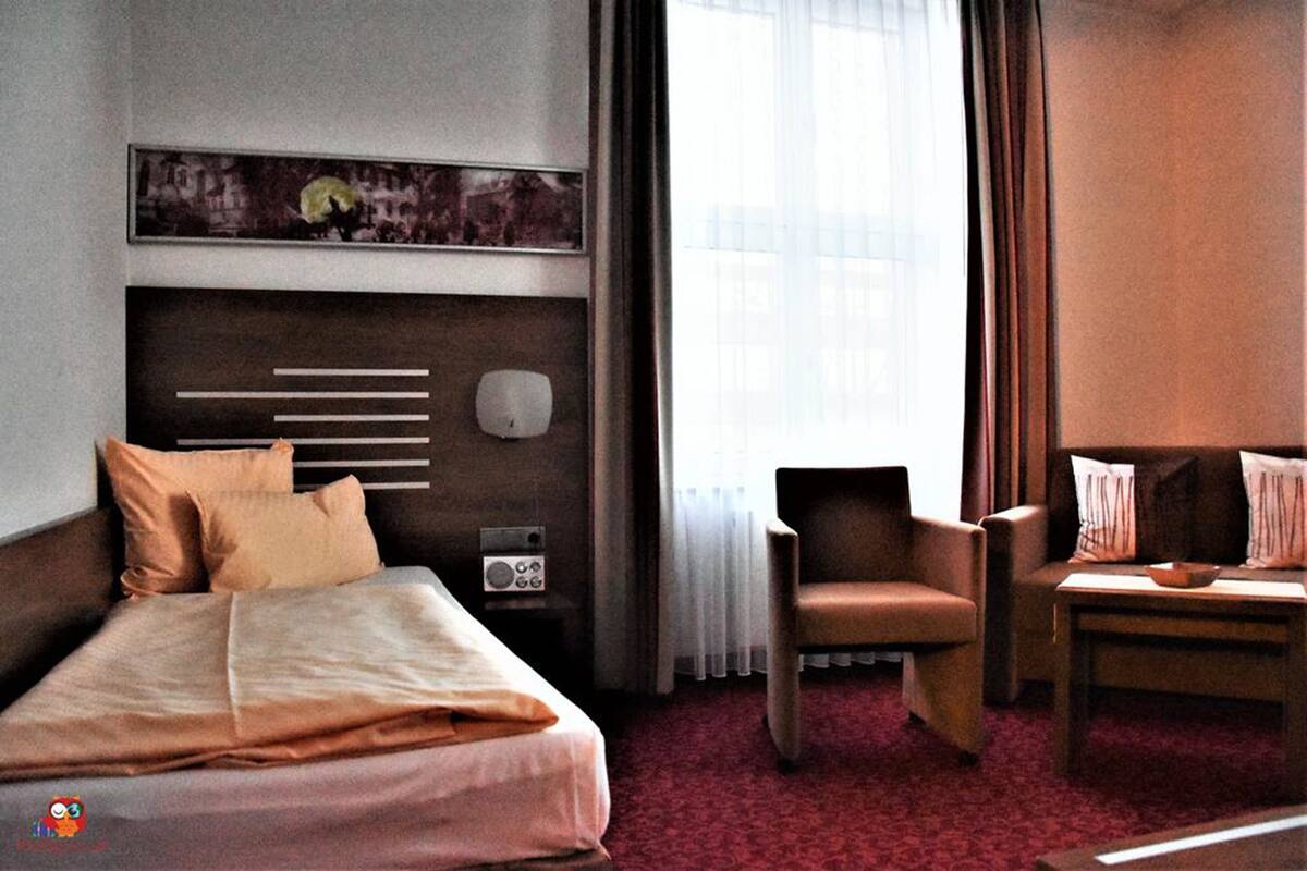 Una habitación de hotel con cama, una silla, una mesita, un radio y un cuadro en la pared.