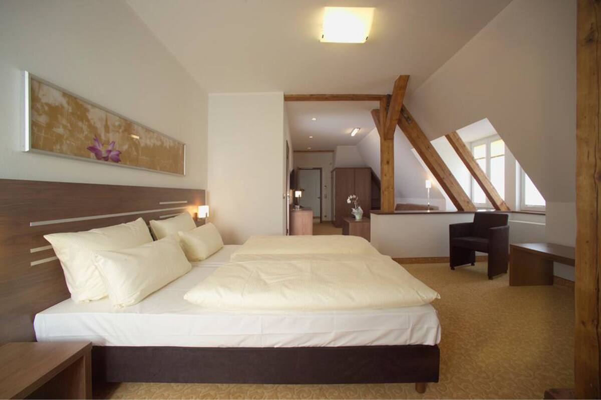Un dormitorio con una cama grande, cabecera de madera, una mesita de noche, un escritorio y una silla. Se ve una ventana con vistas al exterior.