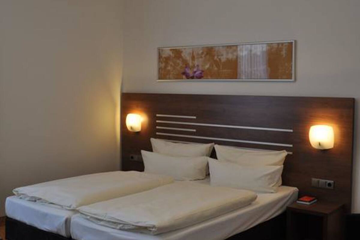 Una cama doble con dos almohadas, cabecera con lámpara, mesita de noche pequeña y una obra de arte colgada en la pared.