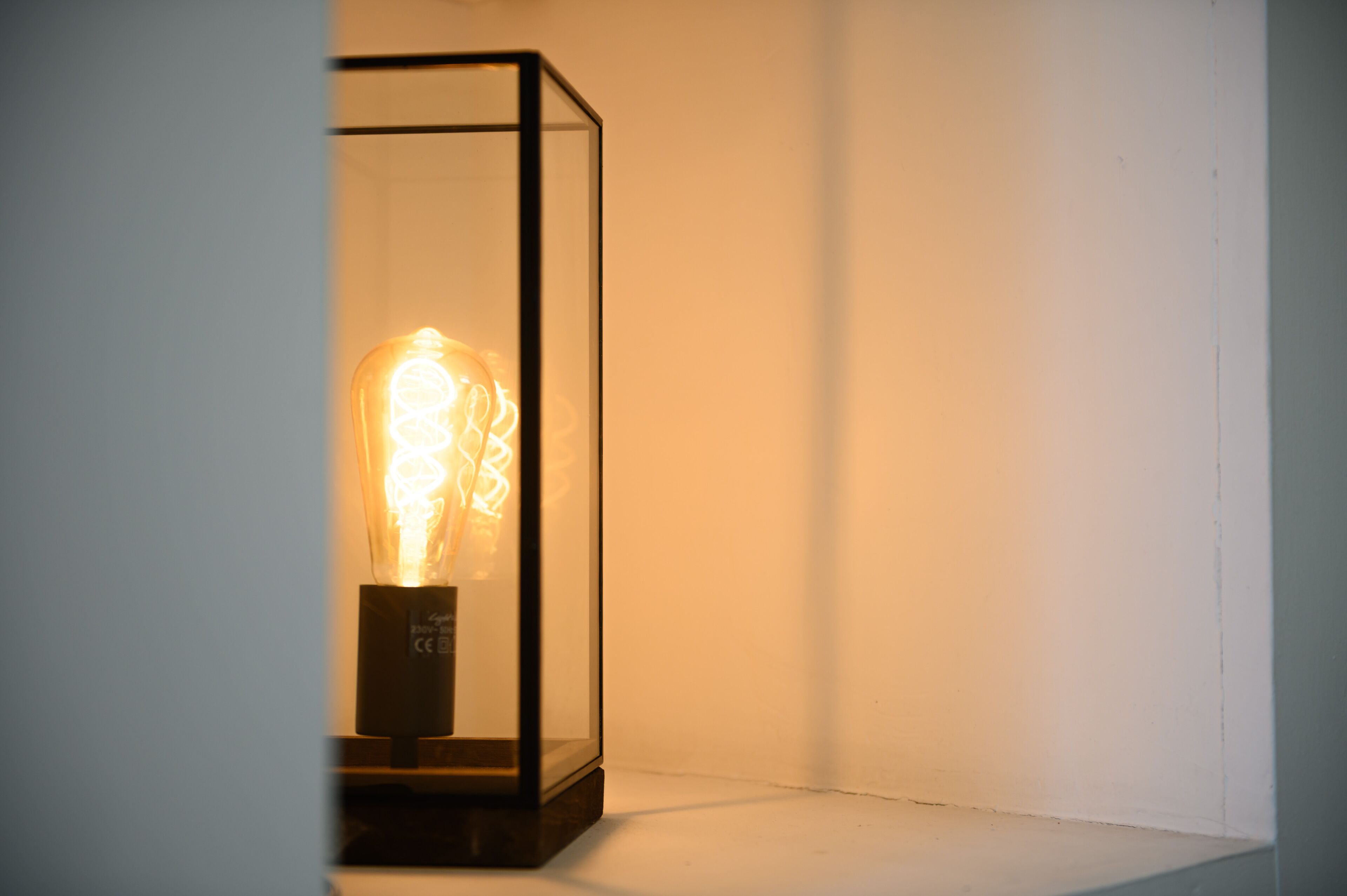 A light bulb encased in a clear glass box with a black base, placed against a plain wall.