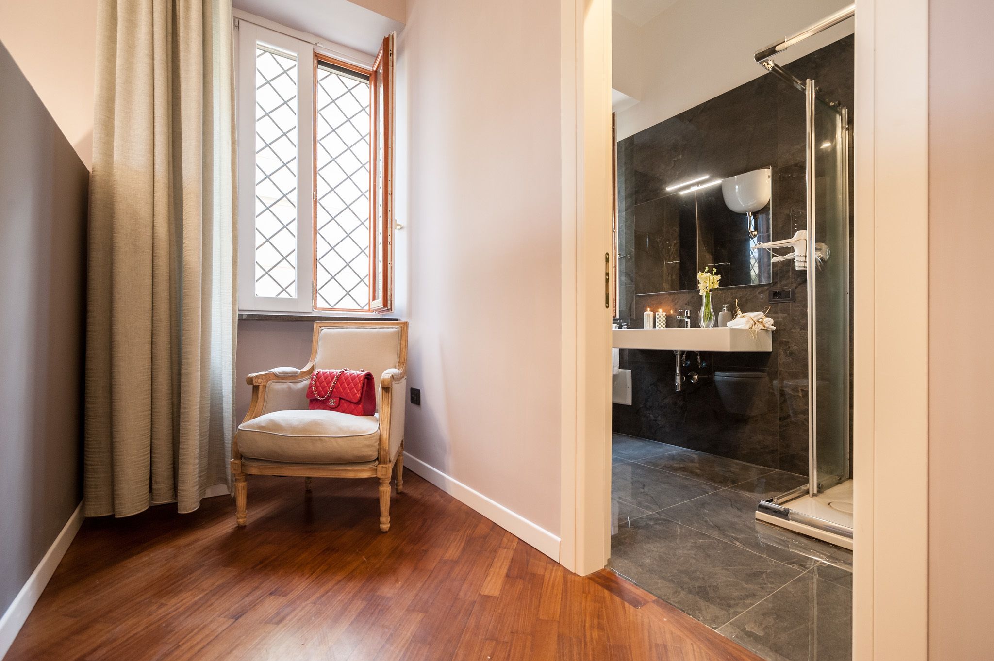 A hallway leading to a bathroom with a shower, a chair, and a window with curtains.