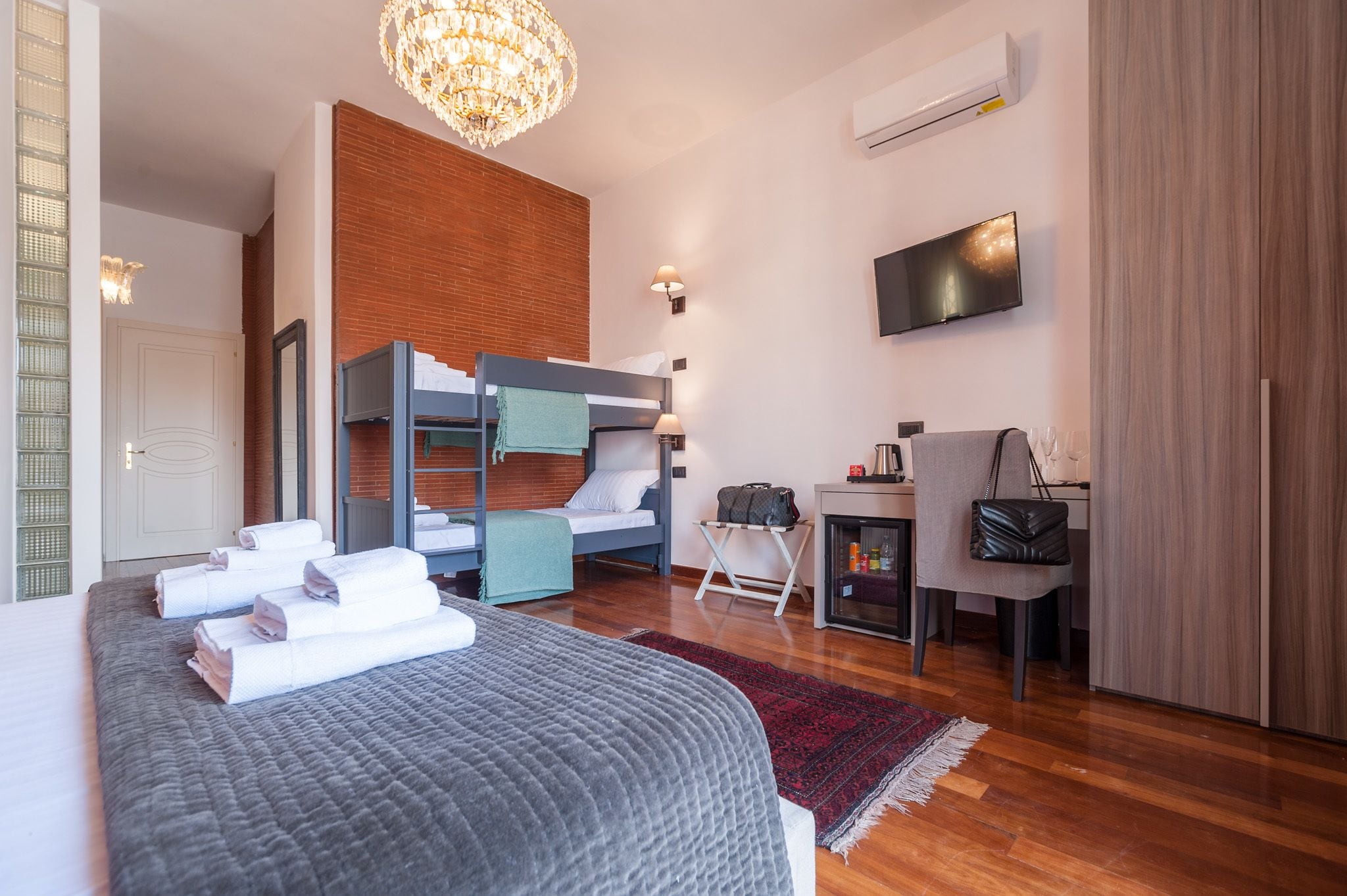 A modern hotel room with a bunk bed, a desk with a chair, a television, and a minibar.