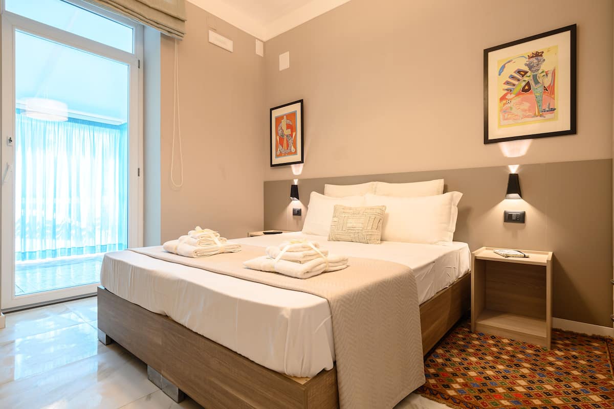 Quarto de hotel moderno com uma cama grande, uma pequena mesa de cabeceira, um criado-mudo, uma janela com cortinas e duas luminárias fixadas na parede.