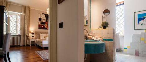 Habitación de hotel con piso de madera, cama, mesita de noche, cómoda, baño con lavamanos y espejo, y ventana con cortinas.