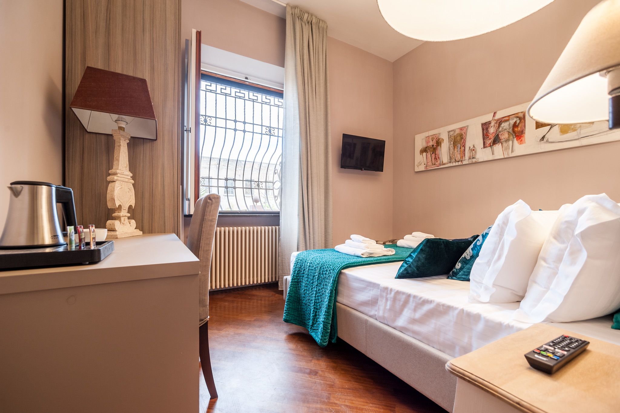 Et hotelværelse med seng, skrivebord med lampe, fjernsyn og billedkunst på væggen.