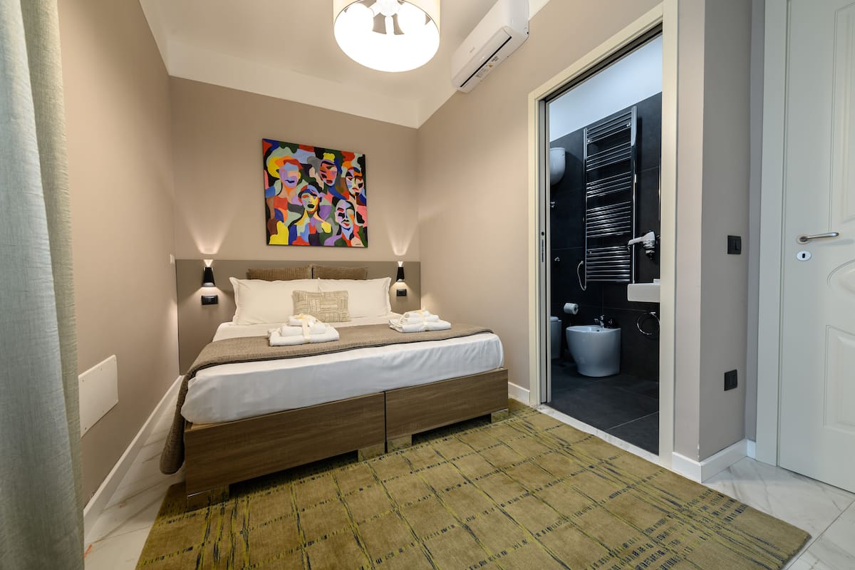 Quarto de hotel moderno com cama, pintura abstrata colorida, banheiro com chuveiro e porta que dá para o exterior.
