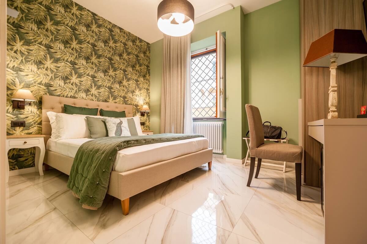 Quarto com cama, mesas de cabeceira, uma cadeira, escrivaninha e janela com cortinas.