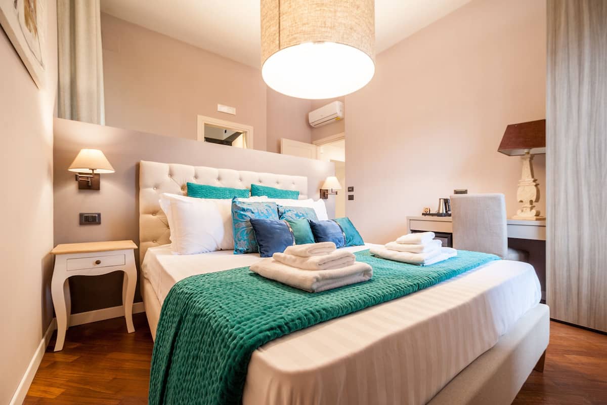 Cama bem arrumada com um cobertor verde, mesa de cabeceira com abajur e escrivaninha com luminária.
