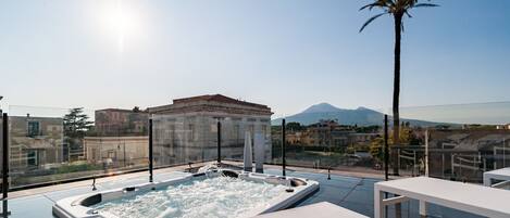 A rooftop terrace with a hot tub, lounge chairs, and a view of buildings and mountains.