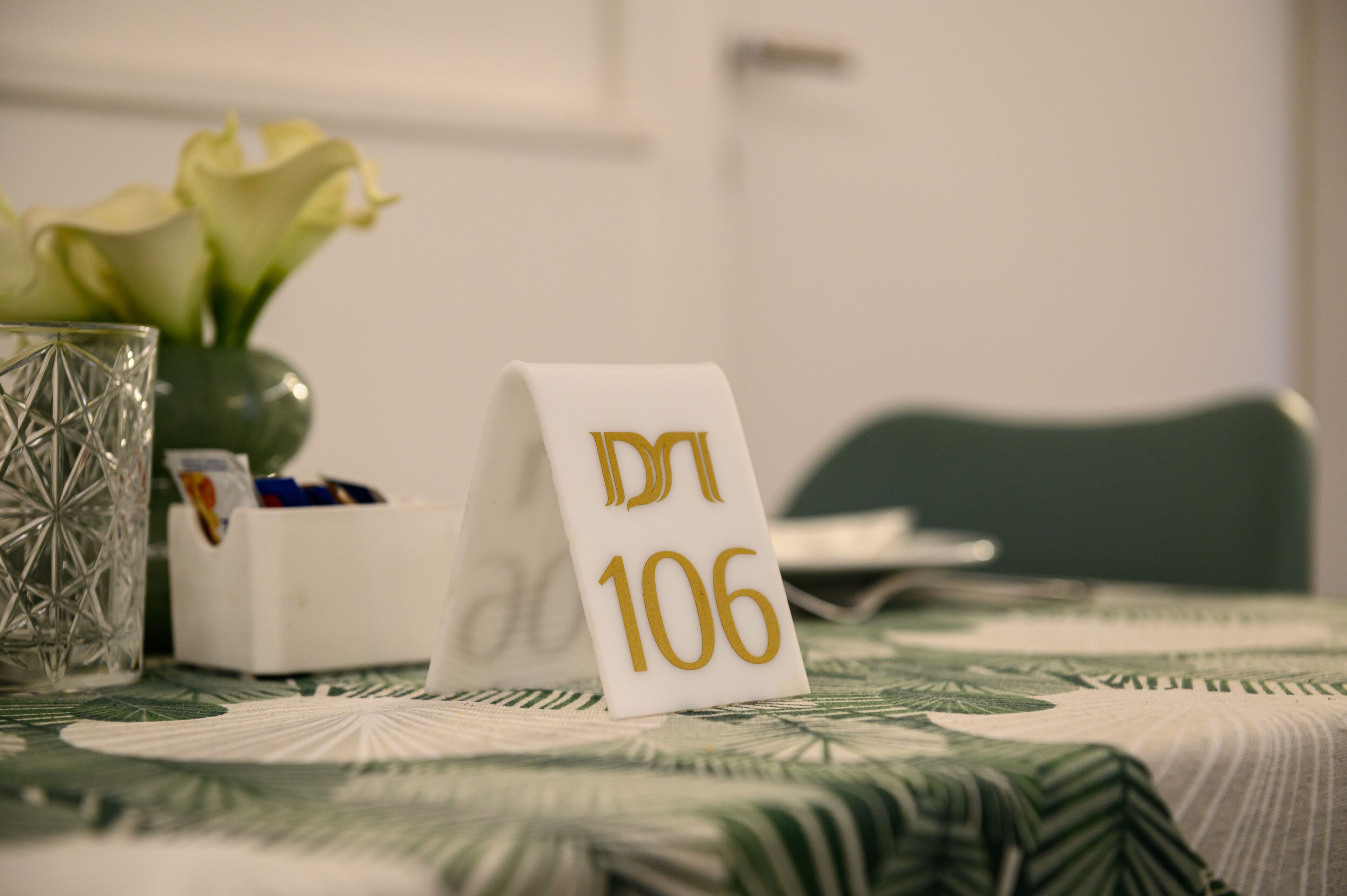 Une table avec un set de table, un vase en verre contenant des fleurs, une pancarte blanche avec du texte en hébreu et une boîte blanche contenant des stylos.