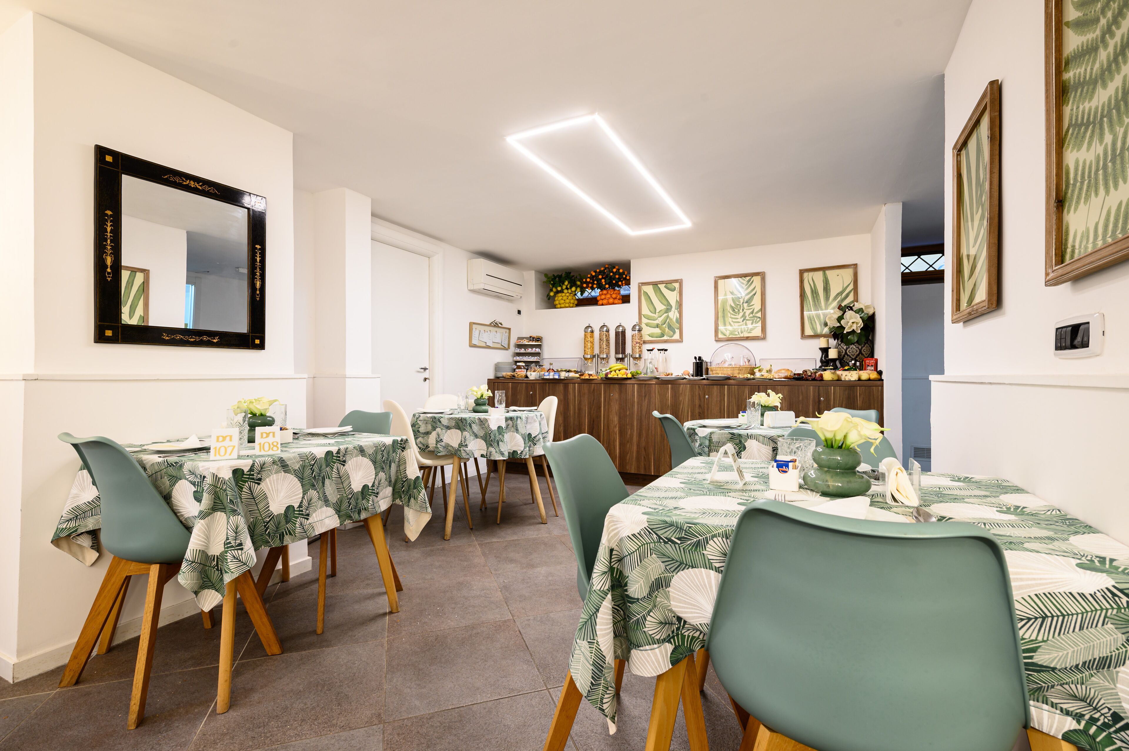 Un’area pranzo con tavoli apparecchiati per quattro, ognuno con una sedia verde e una tovaglia con motivo a foglia. Sullo sfondo, si distingue una zona bar con diversi oggetti esposti.