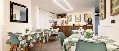 A dining area with tables set for four, each with a green chair and a tablecloth with a leaf pattern. There is a bar area in the background with various items on display.