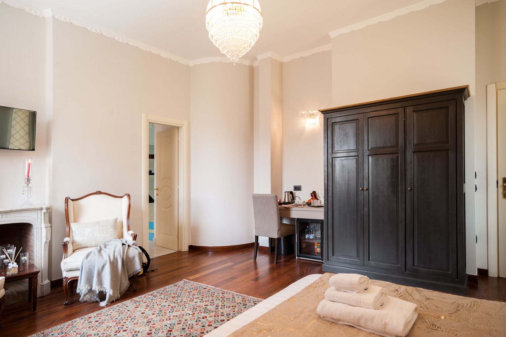 A room with a wooden floor, a large dark wooden wardrobe, a desk with a chair, a fireplace, and a chandelier.