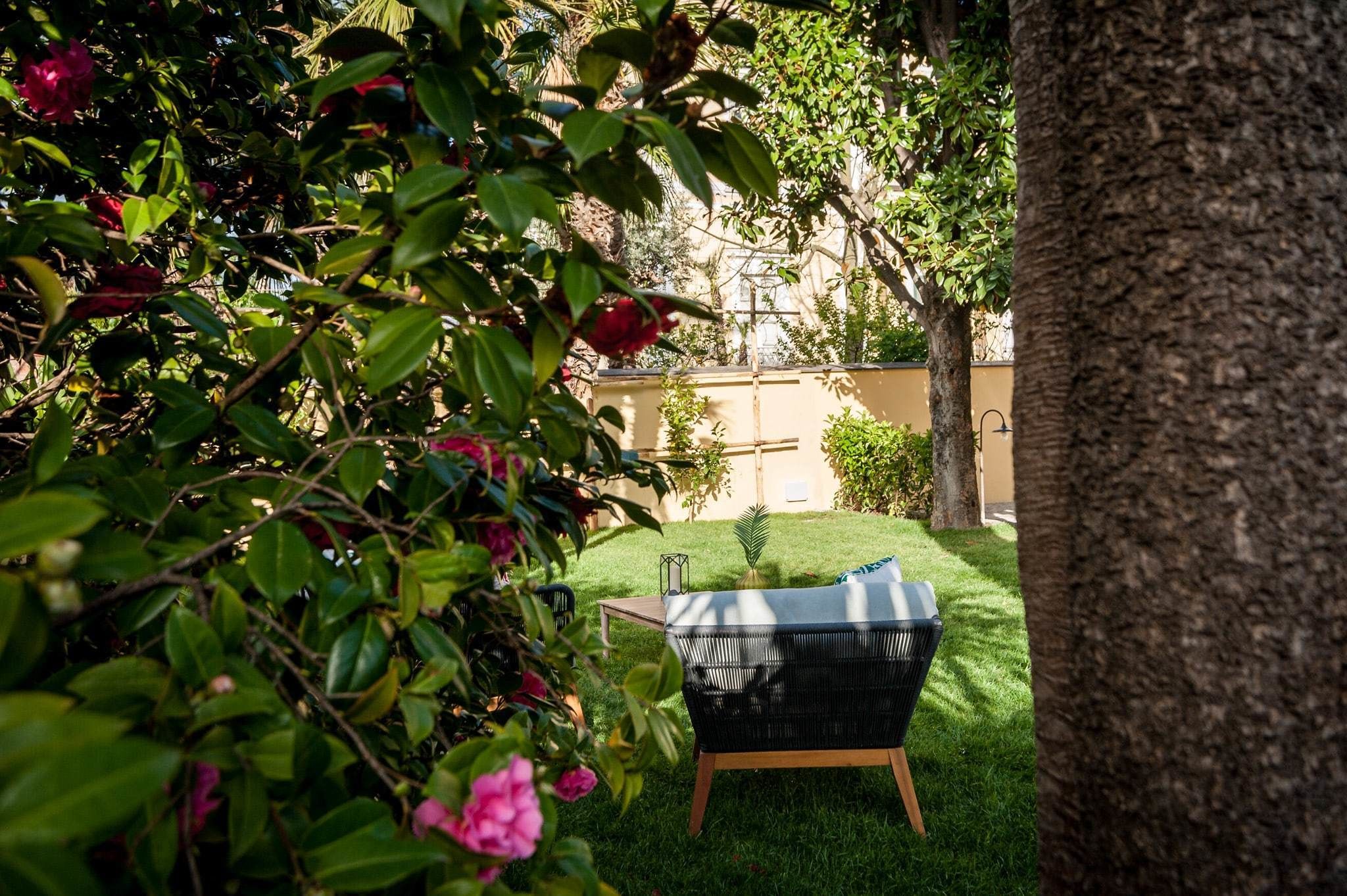 A garden with a wicker chair and a small table, surrounded by lush greenery and a tree.