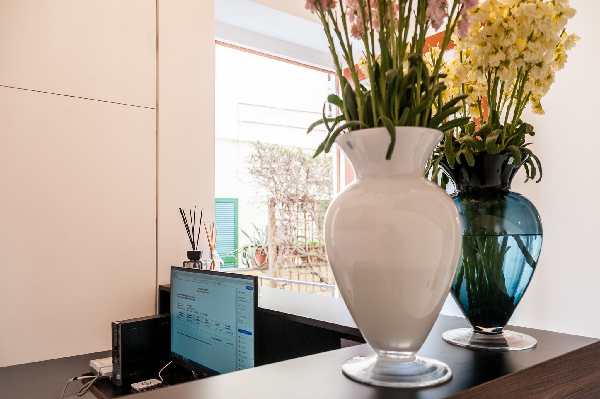 Un bureau avec un écran d’ordinateur affichant une page web, un vase blanc avec des fleurs et un vase bleu avec des fleurs.