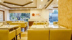 Interno di un ristorante con sedute e tavoli gialli, un cartello con la scritta "QUBE café" e vista su del verde all'esterno.