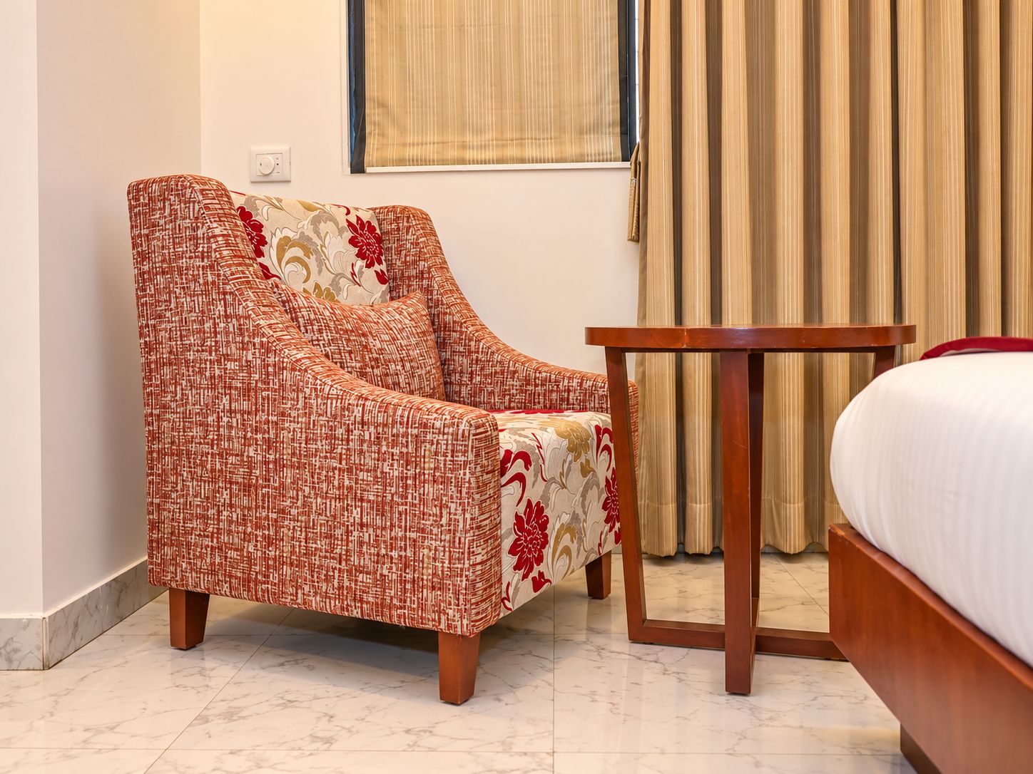 A hotel room with a single bed, a wooden nightstand, a red and beige armchair, and a window with curtains.