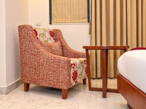 Habitación de hotel con una cama individual, una mesita de noche de madera, un sillón rojo y color beige, y una ventana con cortinas.