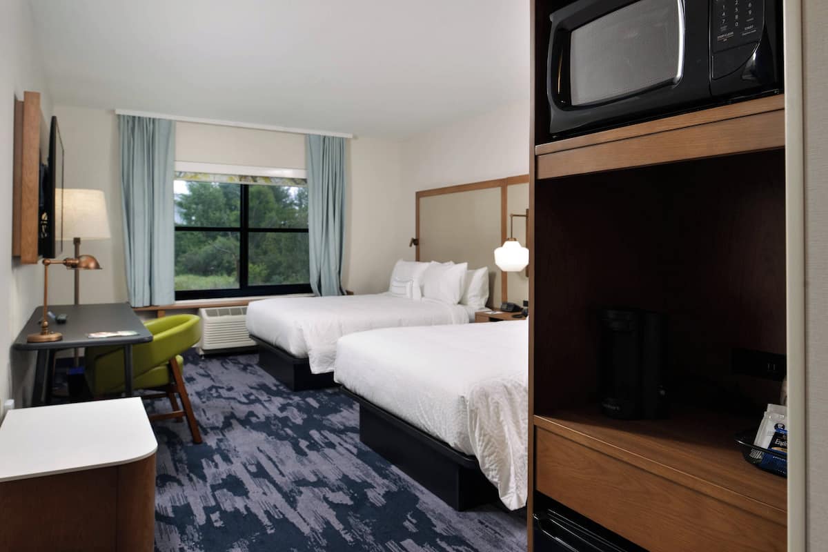 A hotel room with two beds, a desk, a chair, a microwave, and a window with curtains.