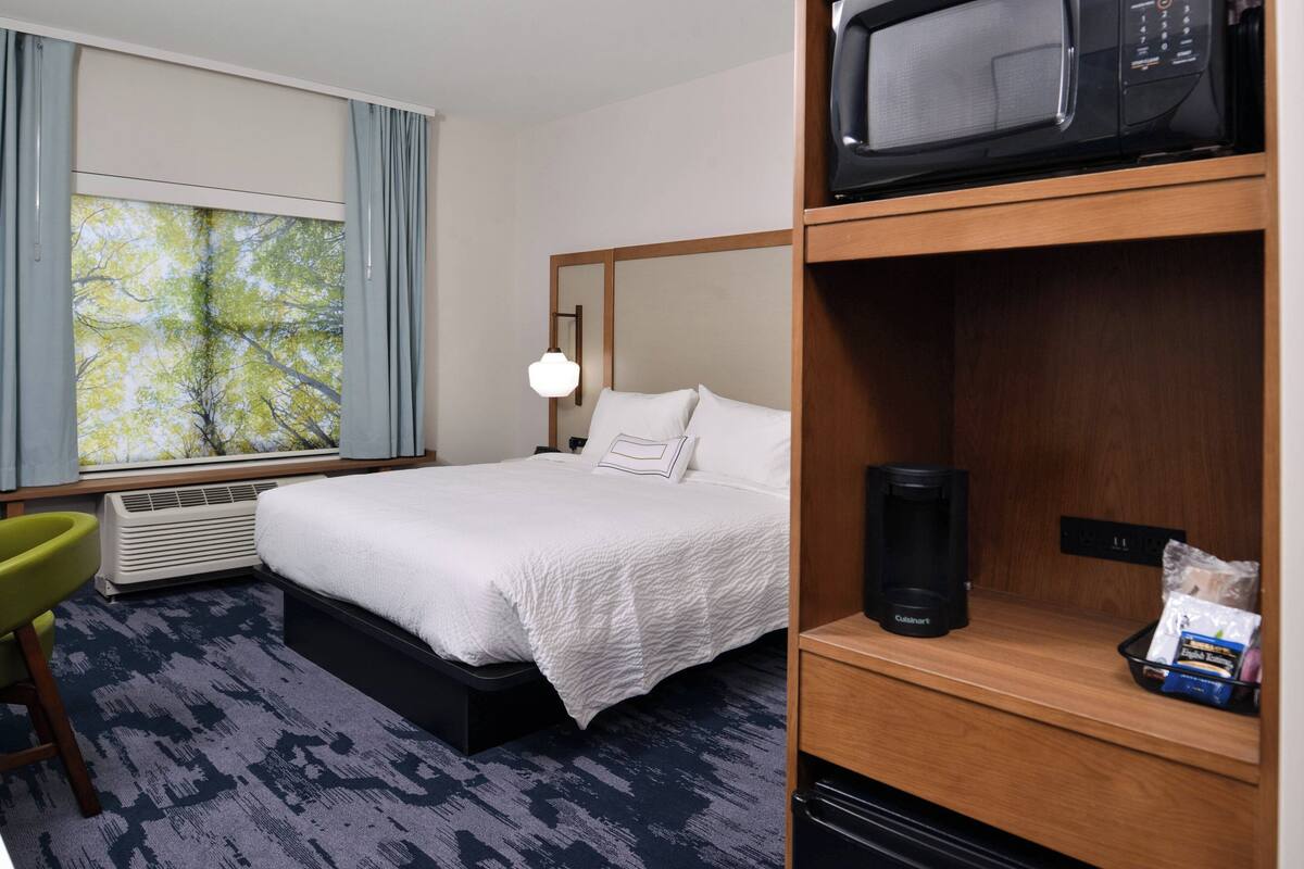 A hotel room with a bed, a window with curtains, a microwave, a coffee maker, and a small table with a bag of snacks.
