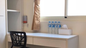 Un escritorio blanco con una silla negra, una ventana con cortinas color beige y tres botellas de agua en el alféizar.
