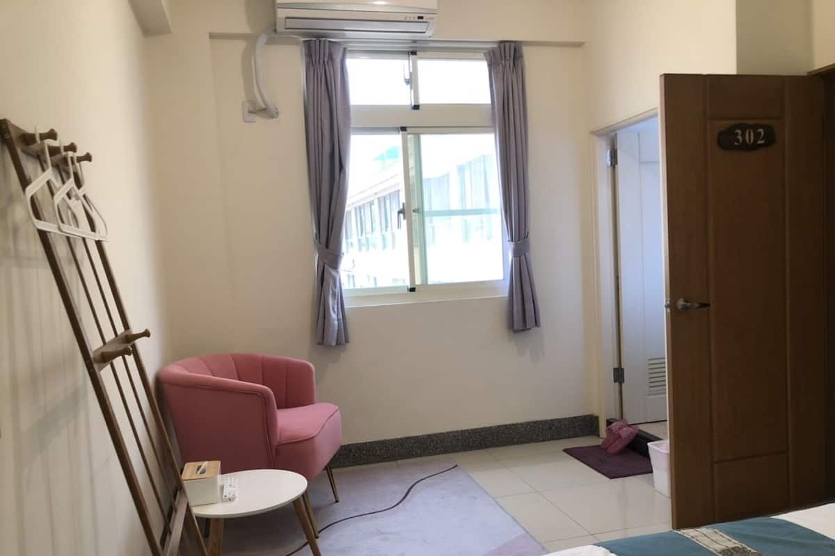 A hotel room with a bed, a pink chair, a small table, a window with curtains, and a door with a number.