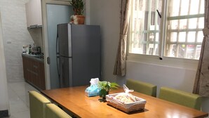 Un comedor con una mesa de madera, sillas con cojines verdes, un refrigerador y una ventana con cortinas.