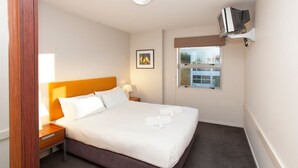 A neatly made bed with white linens and a brown headboard, a bedside table with a lamp, a framed picture on the wall, and a window with a view of buildings outside. - Apartment 25 (Wanaka)