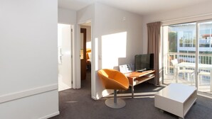 A modern hotel room with a large sliding glass door leading to a balcony, a flat-screen TV, a white desk, an orange chair, and a white ottoman. - Apartment 25 (Wanaka)
