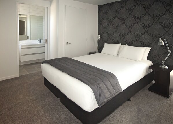 A modern bedroom with a large bed, a nightstand with a lamp, a dresser, and a bathroom visible through an open door.