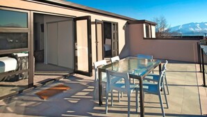 A modern outdoor patio with a glass table and chairs, a barbecue grill, and a view of mountains. - Apartments on Upton (Wanaka)