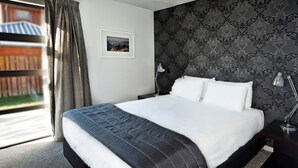 A neatly made bed with white and black bedding, a dark patterned wallpaper, a framed picture on the wall, and a window with curtains. - Apartments on Upton (Wanaka)