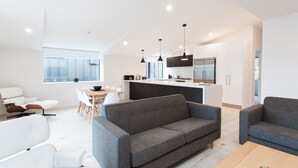 A modern living room with a grey sofa, white armchairs, and a wooden coffee table. There is a kitchen area with a large island, modern appliances, and pendant lighting. - Dungarvon Street Apartments (Wanaka)