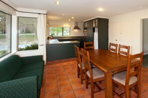 A compact kitchen with a dining table and chairs, a green sofa, and a view of the outdoors through large windows. - Hedditch Hideaway (Wanaka)