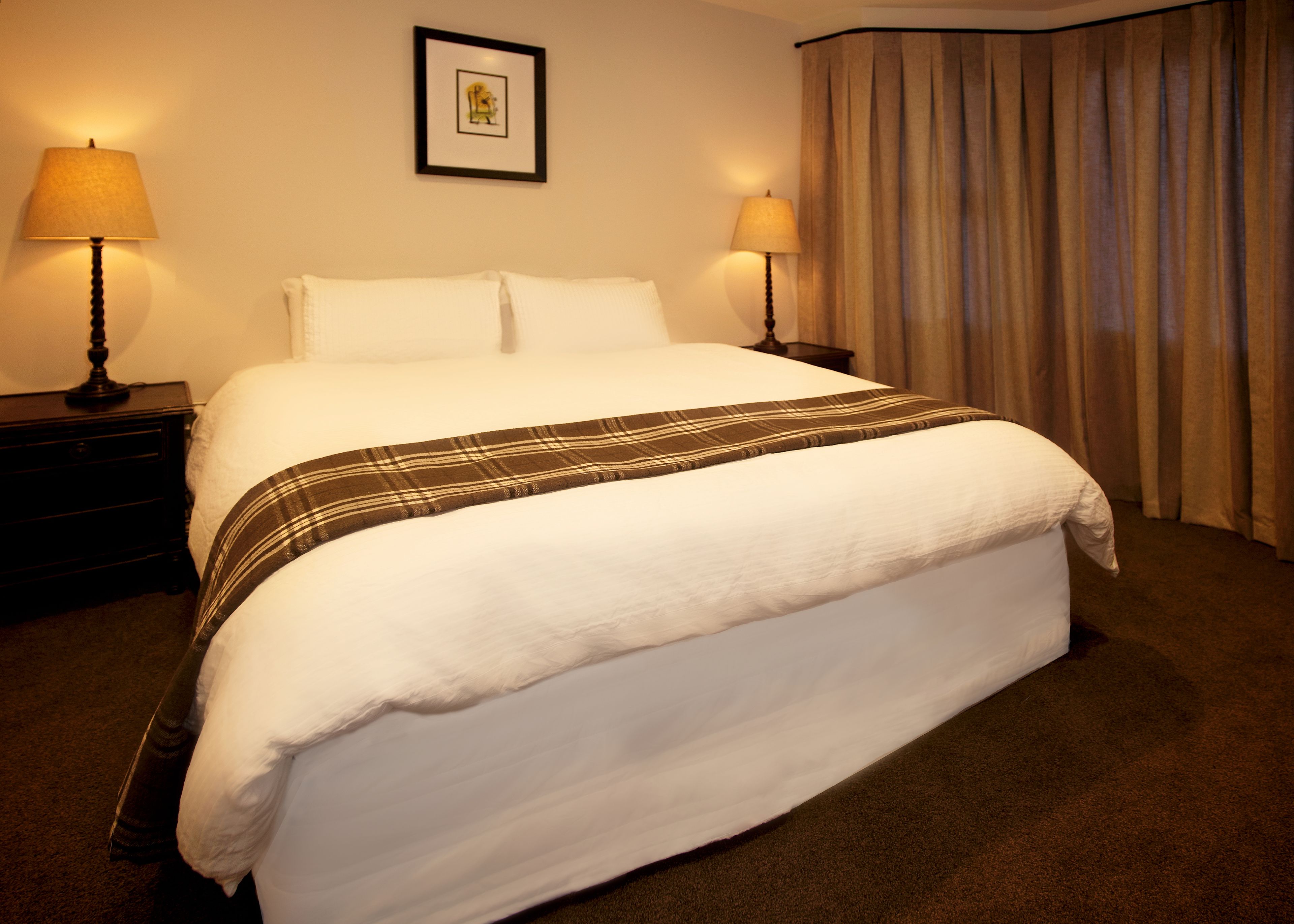 Una cama bien hecha con un plaid, dos lámparas de noche, un cuadro en la pared y una ventana con cortinas.