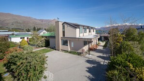 Une maison moderne à deux étages, dotée d’un patio couvert, entourée d’une végétation luxuriante et avec des montagnes en toile de fond.