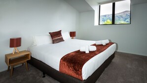 A neatly made bed with white linens and a brown accent pillow, a wooden bedside table with a red lamp, and a view of a mountain landscape through a window. - Rusted Charm (Wanaka)