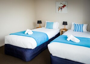 A hotel room with two beds, each with a blue blanket and white pillows, a small table with a lamp, and a wall-mounted artwork. - Villa 62 (Wanaka)