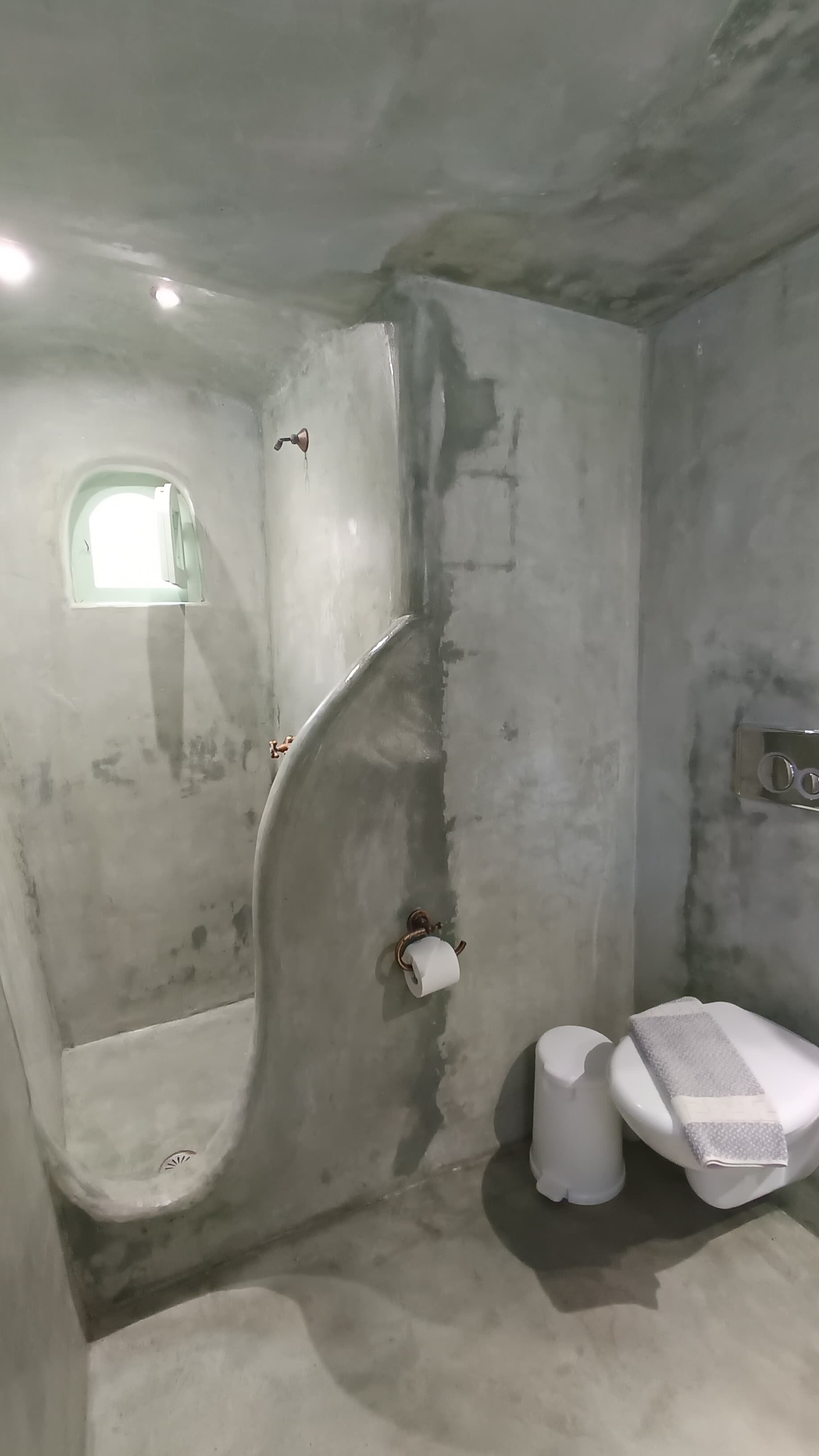 A bathroom with a concrete wall, a small window, a toilet, and a sink.