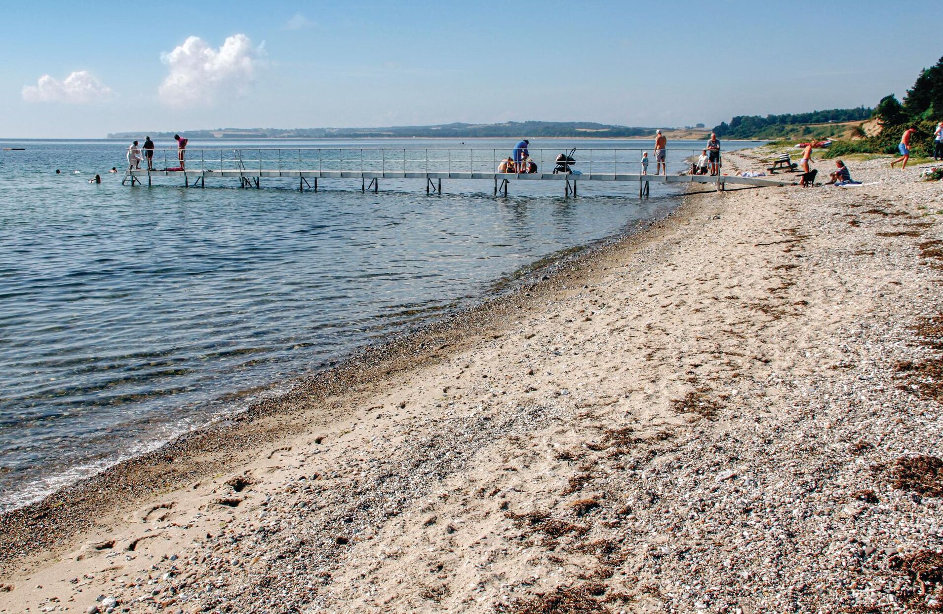 Uma praia com um píer que se estende sobre a água, pessoas caminhando e sentadas no píer, e uma praia arenosa ao fundo.