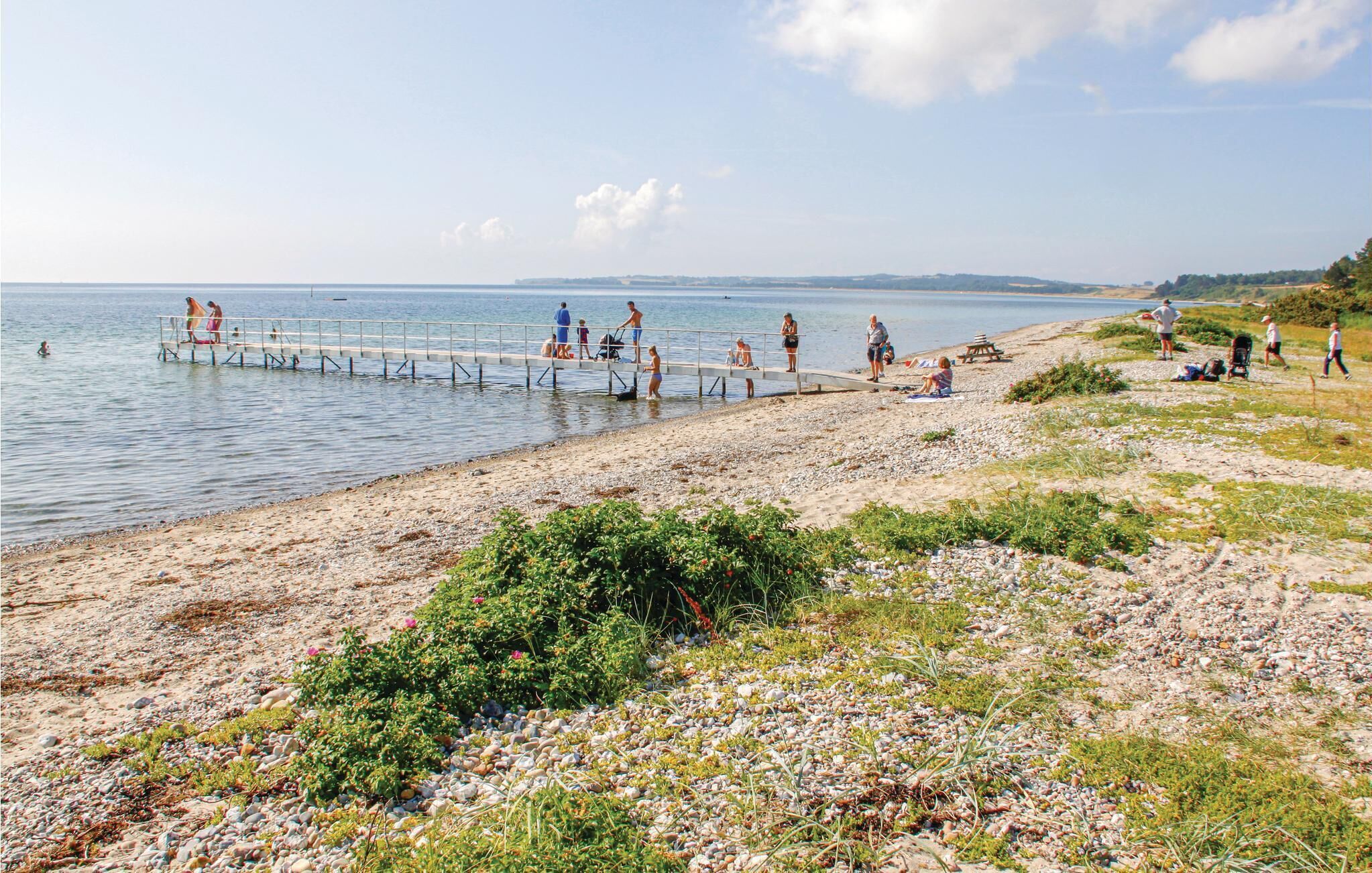 A pebbled beach with a pier extending into calm waters, surrounded by greenery and distant hills.