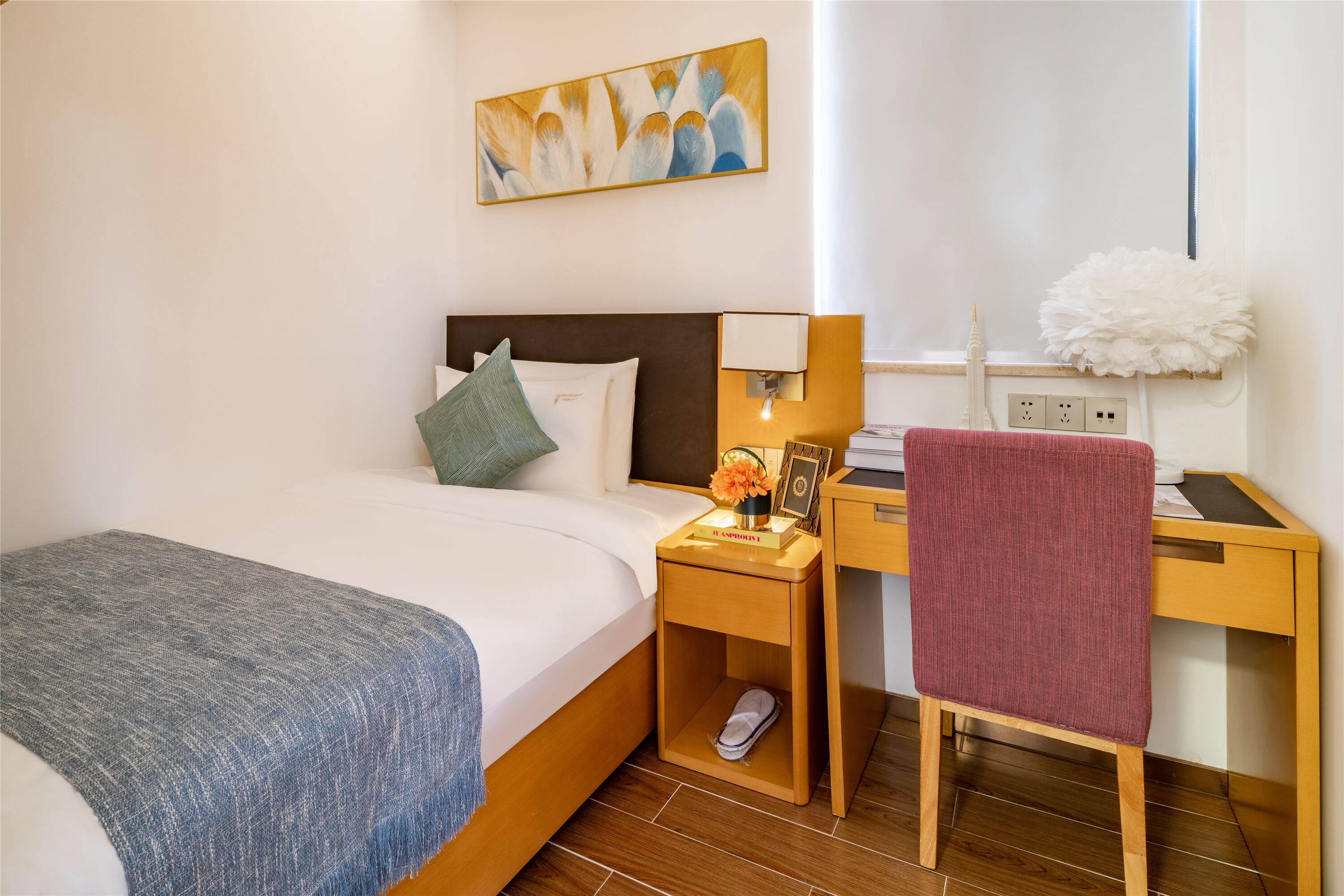 Habitación de hotel con cama, escritorio, silla, mesita de noche, lámpara, un cuadro y un adorno blanco mullido.