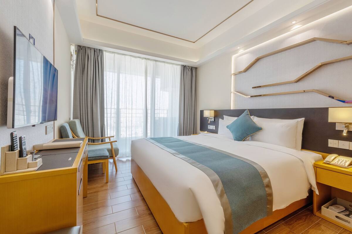Quarto de hotel moderno com uma cama grande, uma escrivaninha com cadeira, uma televisão de tela plana e uma mesa de cabeceira com abajur.