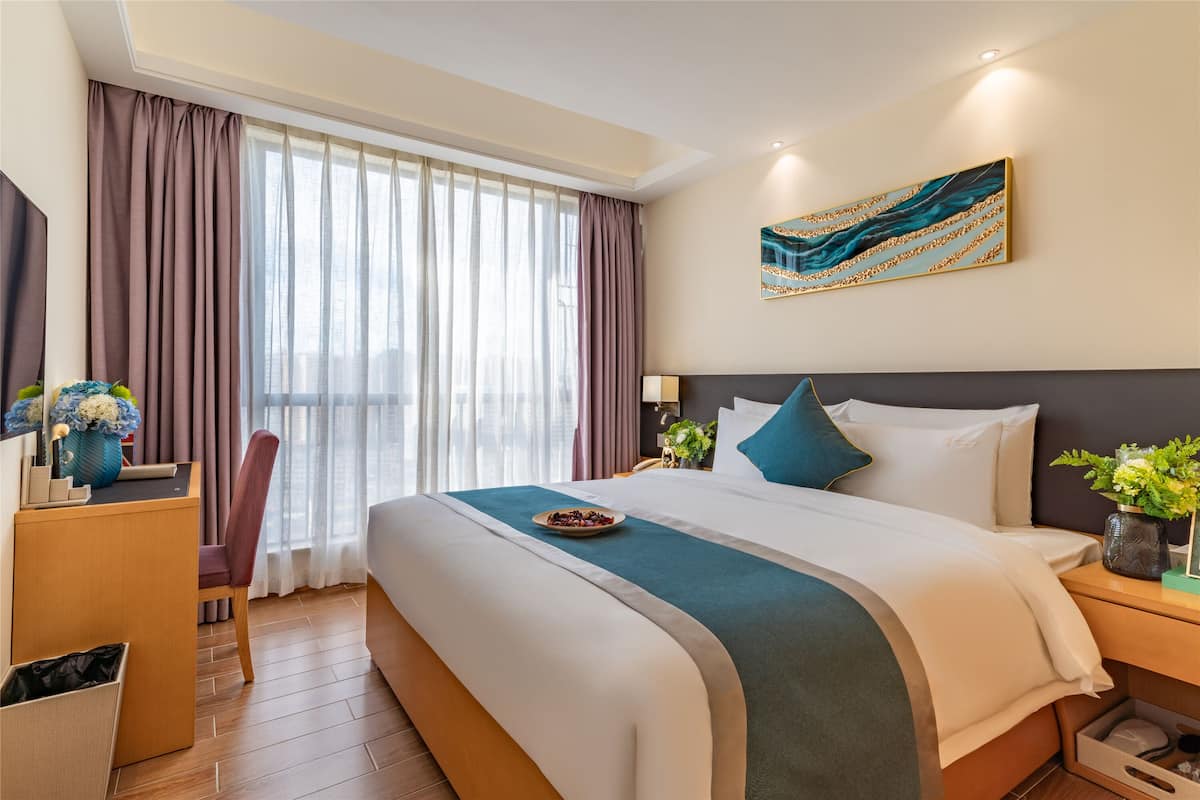 Quarto de hotel com uma cama grande, uma escrivaninha, uma cadeira, uma mesinha de cabeceira com abajur e uma janela com cortinas.