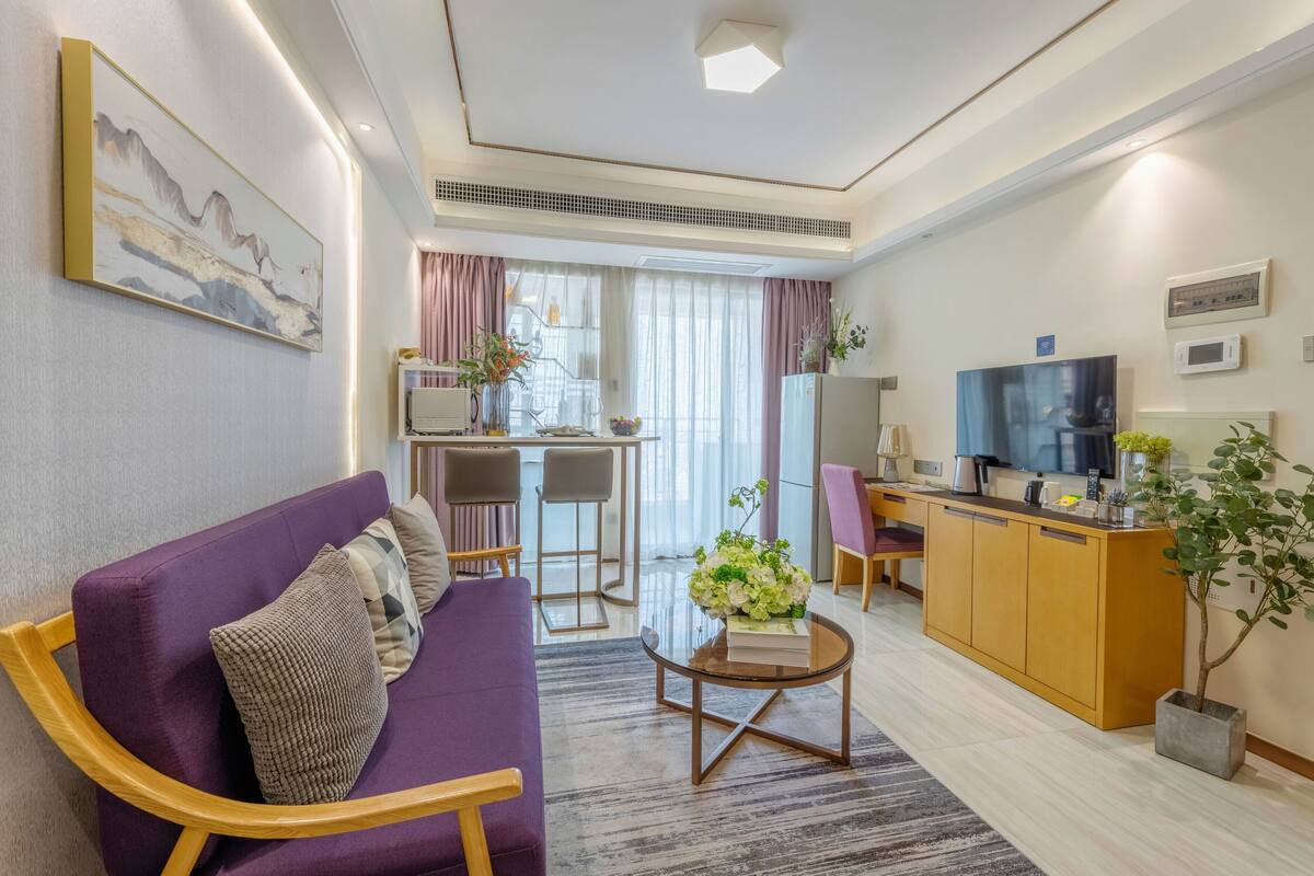 Uma sala de estar moderna com um sofá roxo, uma mesa de centro e uma TV. Há uma área de cozinha com micro-ondas e geladeira.
